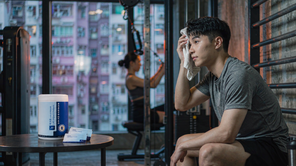 香港健身室內，年輕男子運動後坐在長椅上抹汗，窗外可見密集的舊式住宅大廈